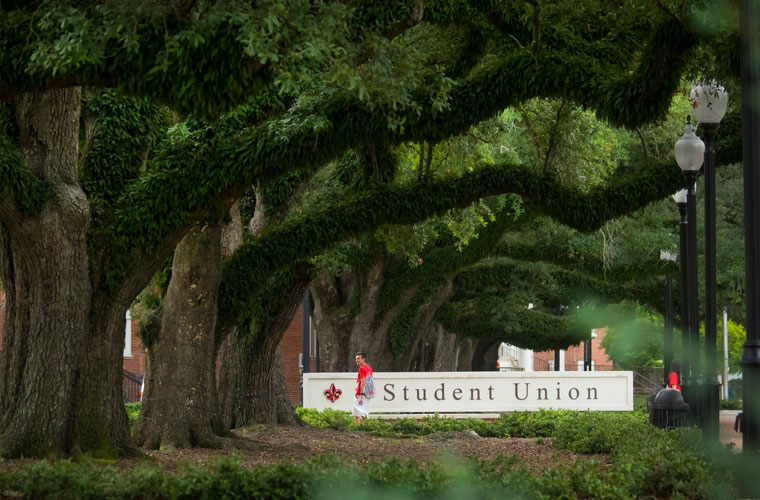 Student walking in front of the University of Louisiana at Lafayette Student Union sign under a canopy of oak tree branches