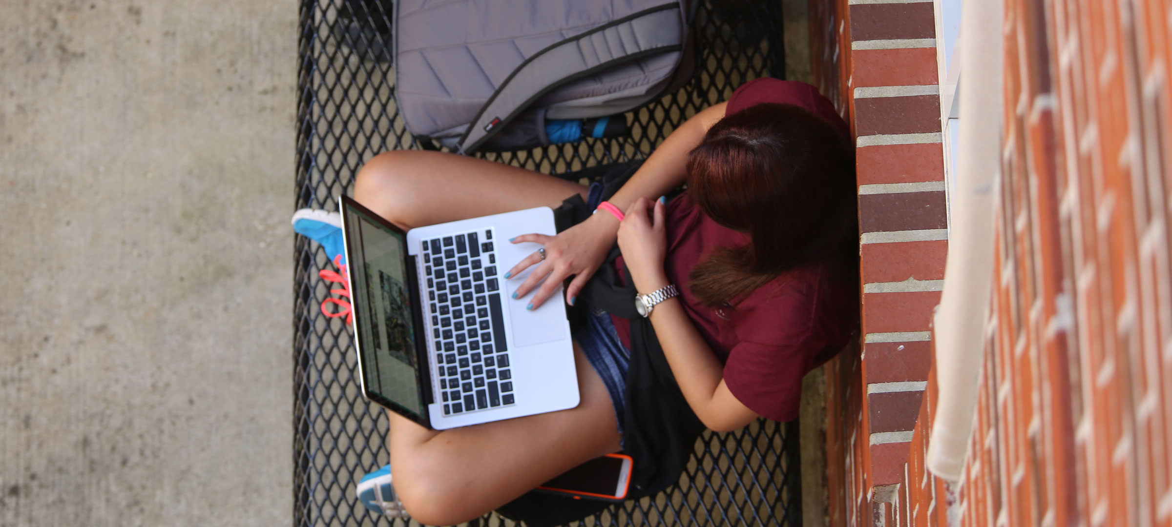 University of Louisiana at Lafayette student sits on campus and looks at her laptop while leaning against a brick wall