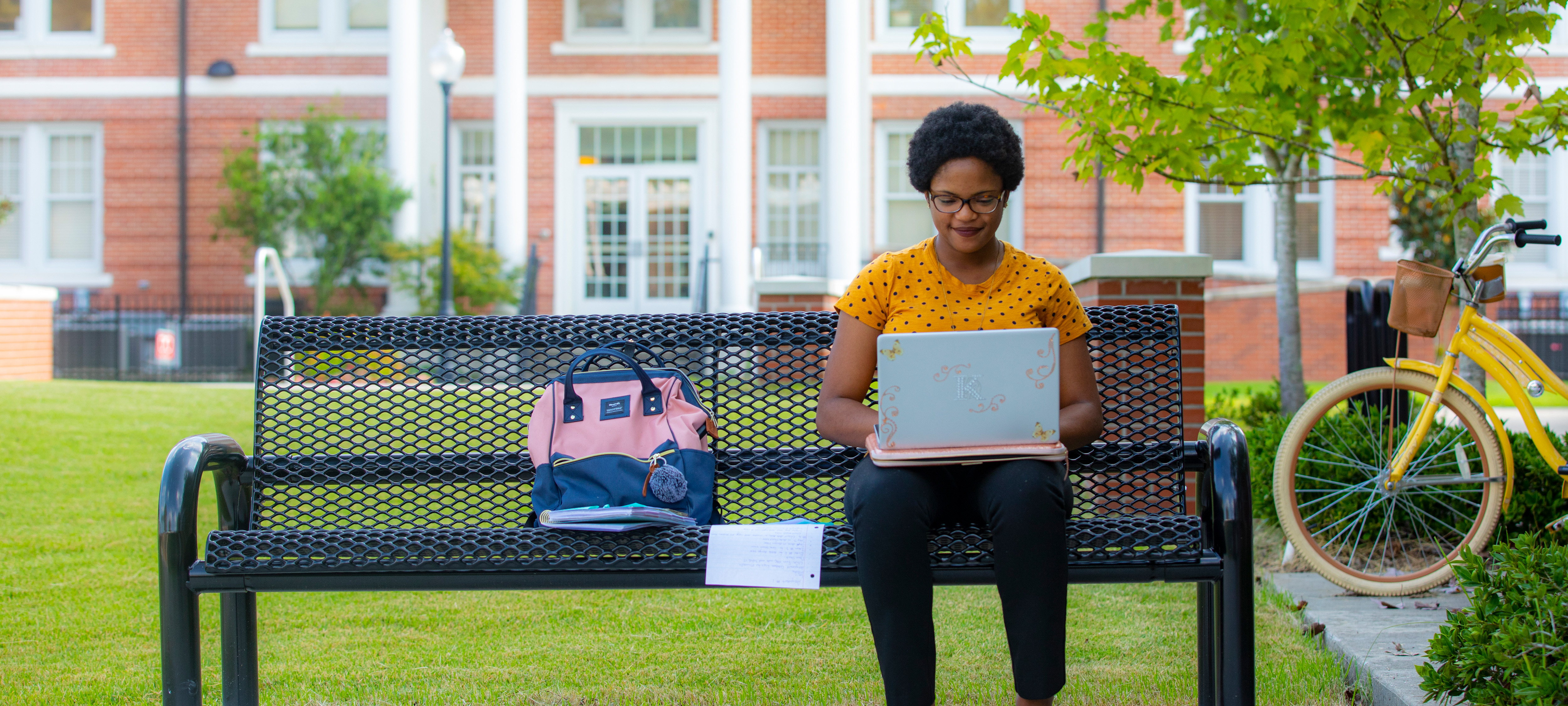 Student sitting on a bench outside using their laptop on UL Lafayette campus.