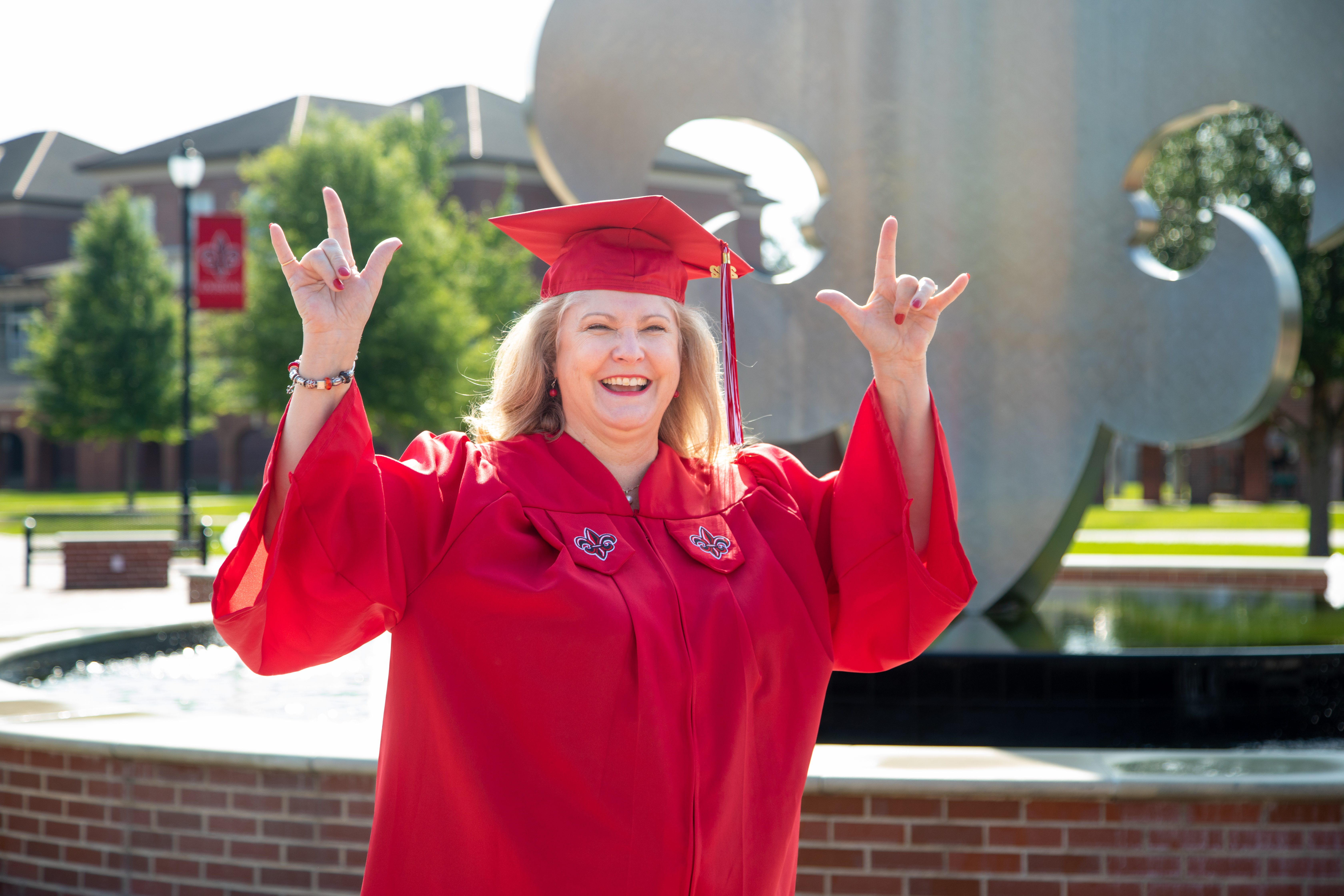 Melanie Fontenot, Bachelor of General Studies Online graduate, is pictured in a red cap and gown in front of the UL Lafayette fleur de lis fountain.