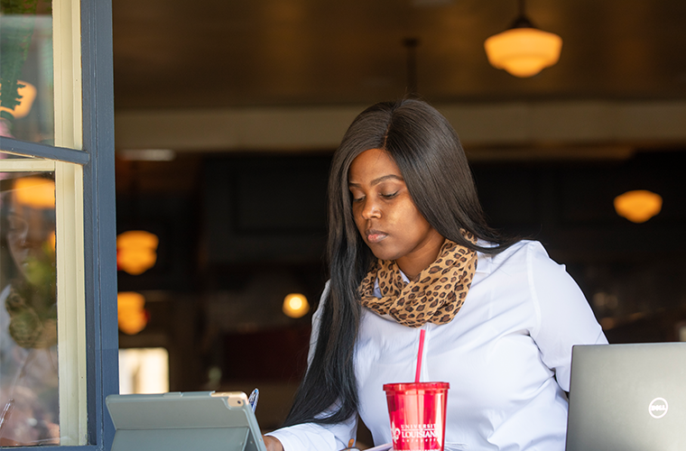 A woman, Kennette Toussaint, peers over a tablet and laptop in an open-air cafe.