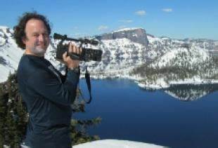Royd Anderson with a camera in his hand over looking water by snowy mountains