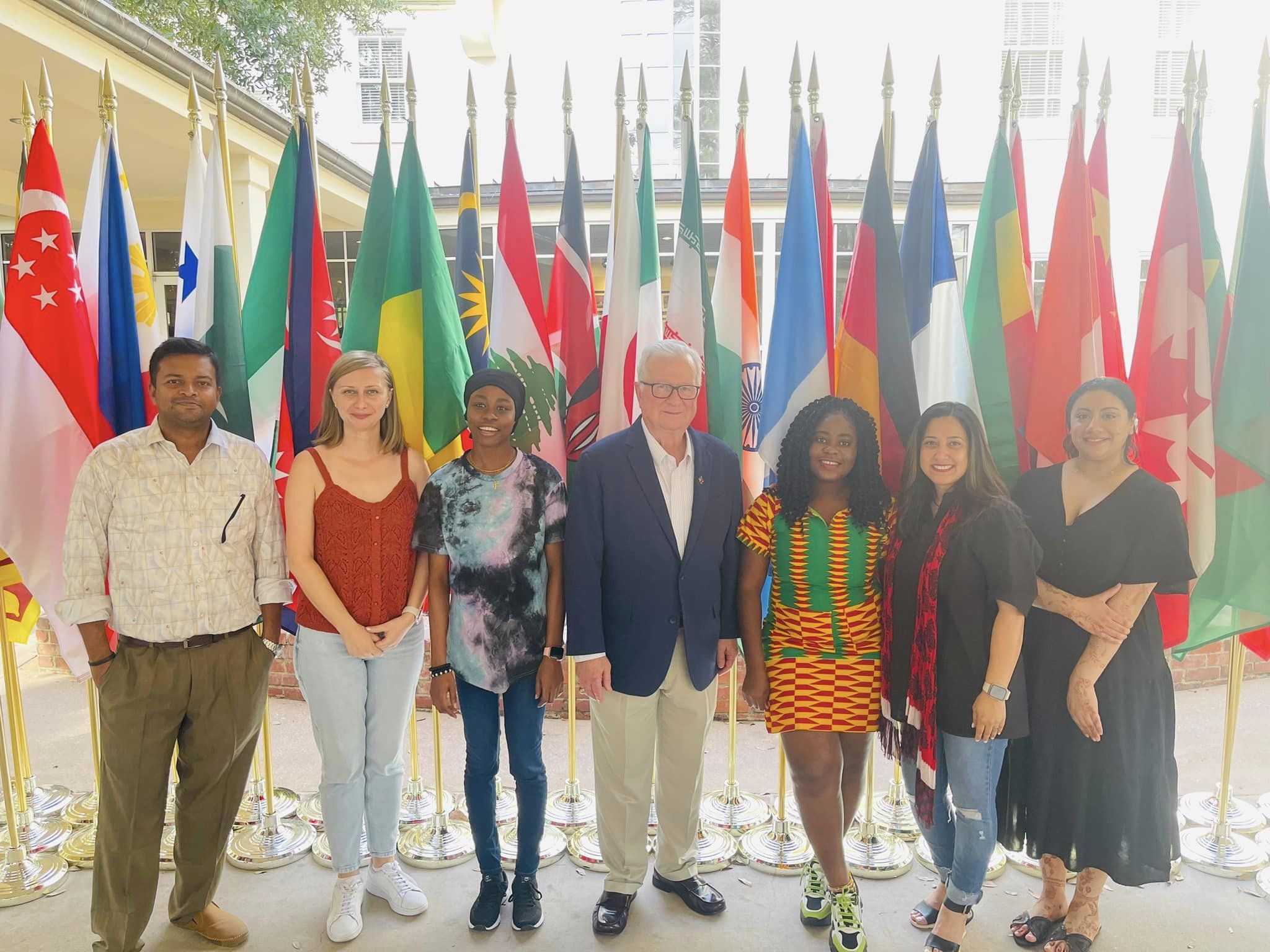 six international students standing with the UL Lafayette President, Dr. Joseph Savoie, in front of many different country flags