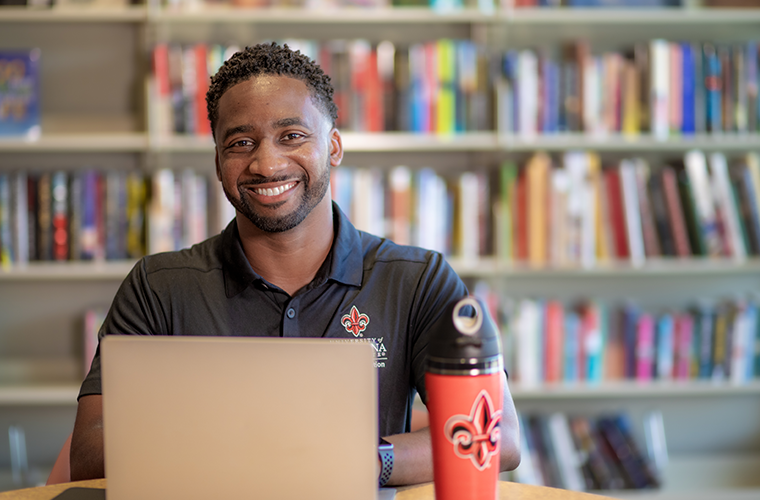 Ryan Owens, MBA, sits at a laptop with a red UL Lafayette water bottle on his left.