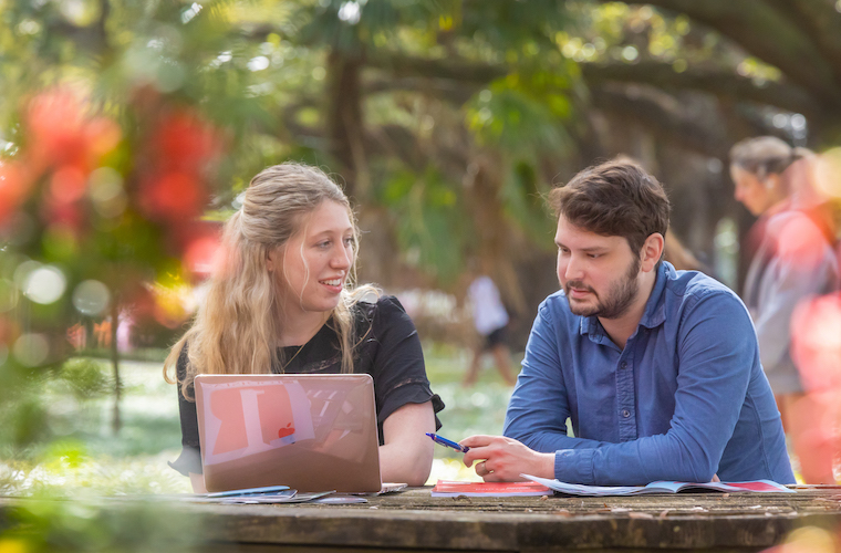 Two students with a laptop and magazines in front of them sitting at an outdoor table on campus.
