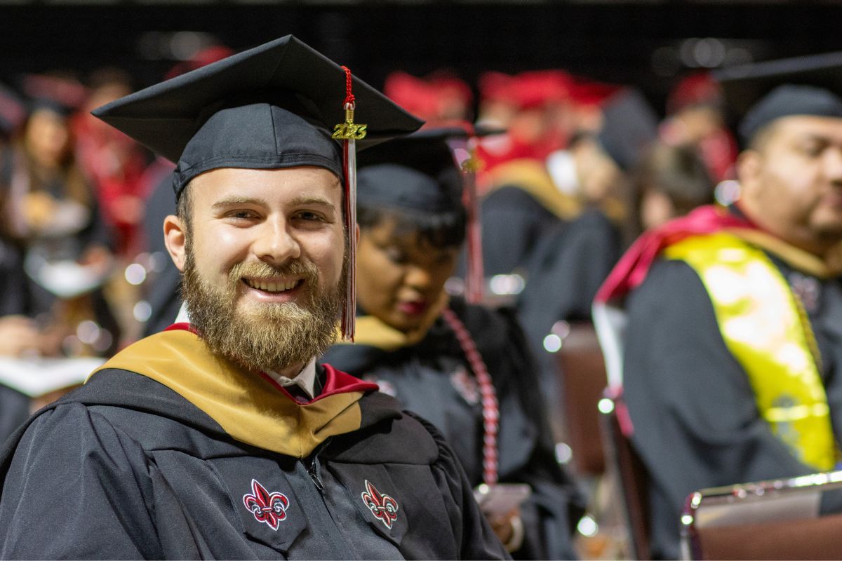 Wearing his master's regalia, Dylan Chesterman sits in his seat at Commencement and smiles at the camera.