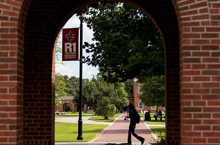A student seen through an archway walking in the UL Lafayette quad by an R1 research flag.