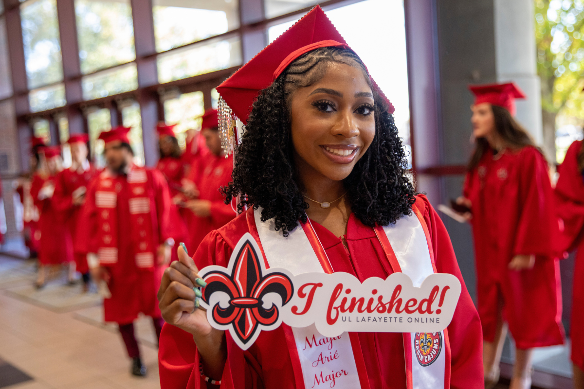 Maya Major smiles at UL Lafayette Commencement ceremony, wearing a red cap and gown and holding a sign that reads: 'I finished!' 