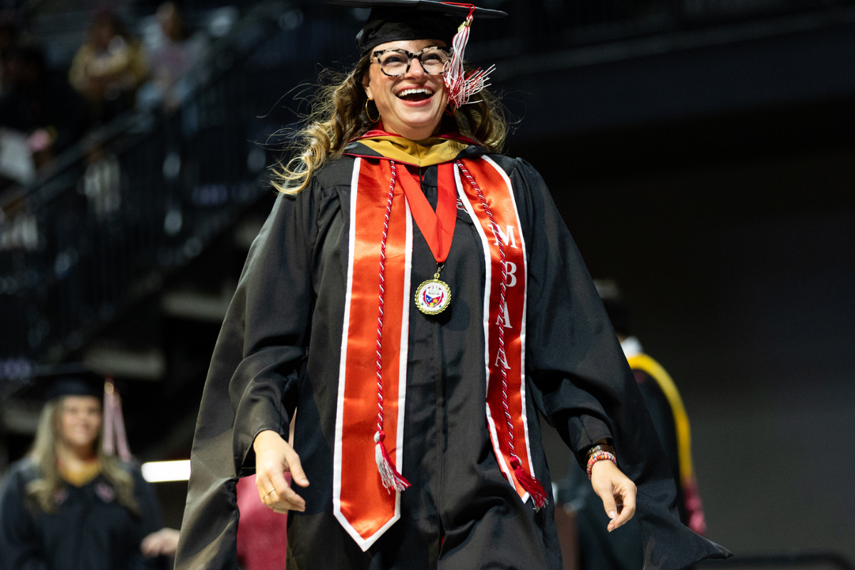 MBA grad Kate Durio crosses the commencement stage wearing a black cap and gown, layered with a red MBA stole and tassles.