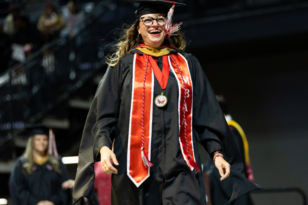 Kate Durio crosses the Commencement stage wearing a black cap and gown layered with a red MBA stole and tassels.