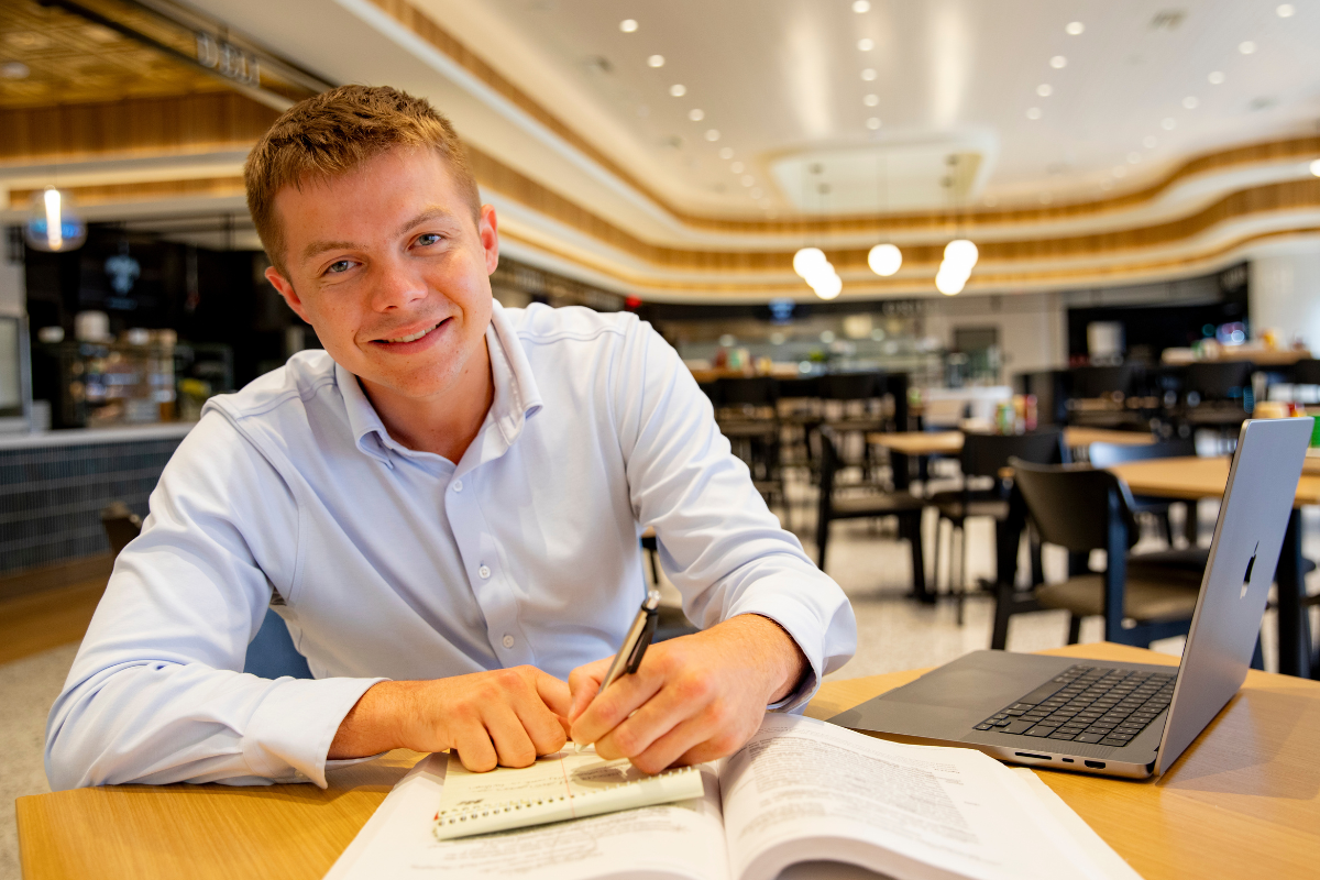 William Fralia, alumnus of UL Lafayette's sport management program, looks up at the camera and smile as he works on his laptop inside of a cafe.