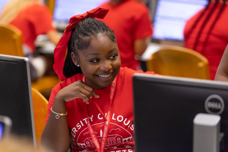 A student in a red Orientation t-shirt sits at a computer smiling in the library computer lab