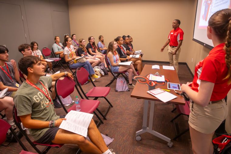 Two Orientation Leaders in red polos stand at the front of a classroom, presenting information to a group of new students