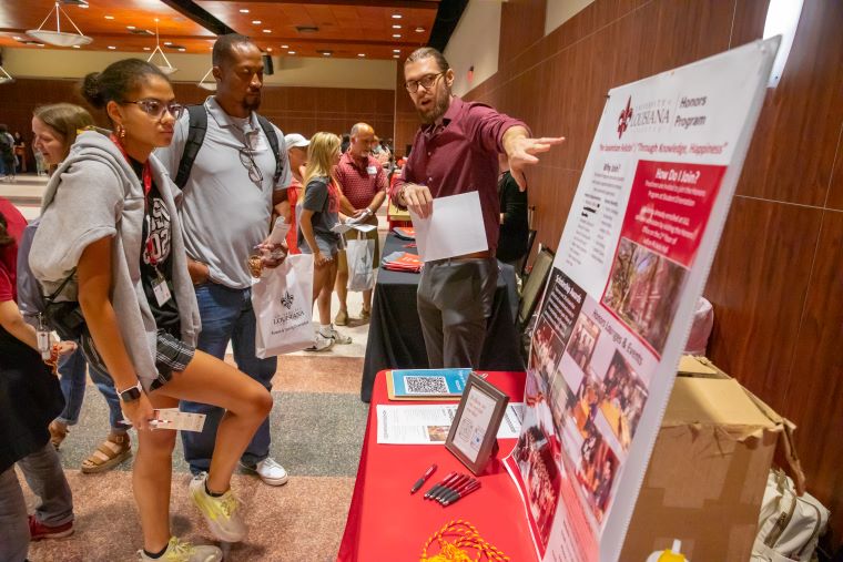 A staff member points to a display board about the Honors Program while speaking to a student and parent
