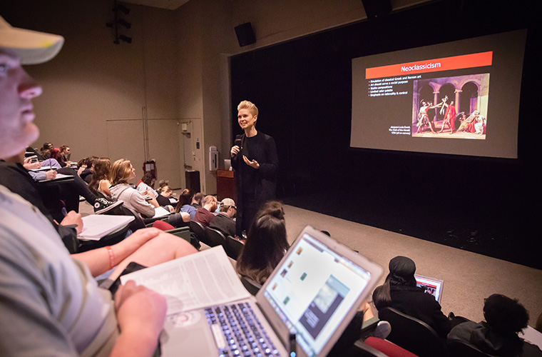 UL Lafayette art history professor lecturing to a large auditorium with a screen behind displaying a slide on Neoclassicism.