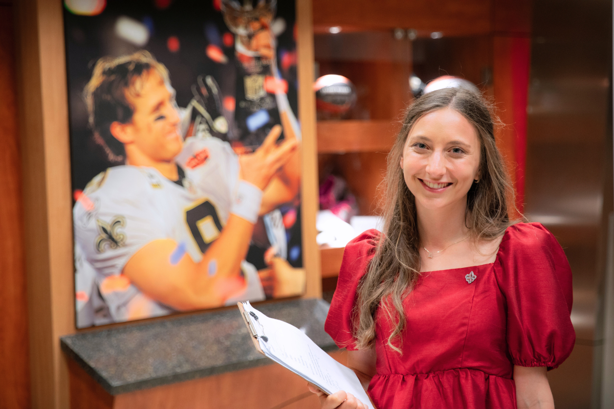 Sport Management program alumni Ashley Livaccari smiles holding a clipboard in front of a portrait of Drew Brees winning the Super Bowl.