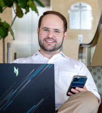 Graduate of UL Lafayette's marketing degree program smiles while multitasking on his work computer and cellphone.