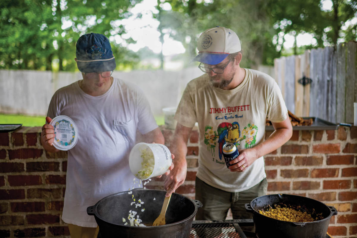 Jacob Gibson and Tanner Dimick pour onions and bell peppers into a black iron pot.