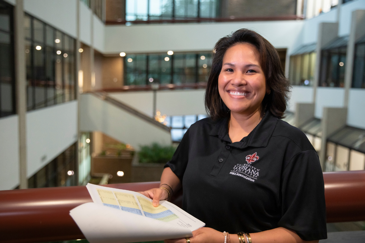 Online MBA in Finance graduate smiles over a data spreadsheet while wearing a black UL Lafayette MBAA polo.