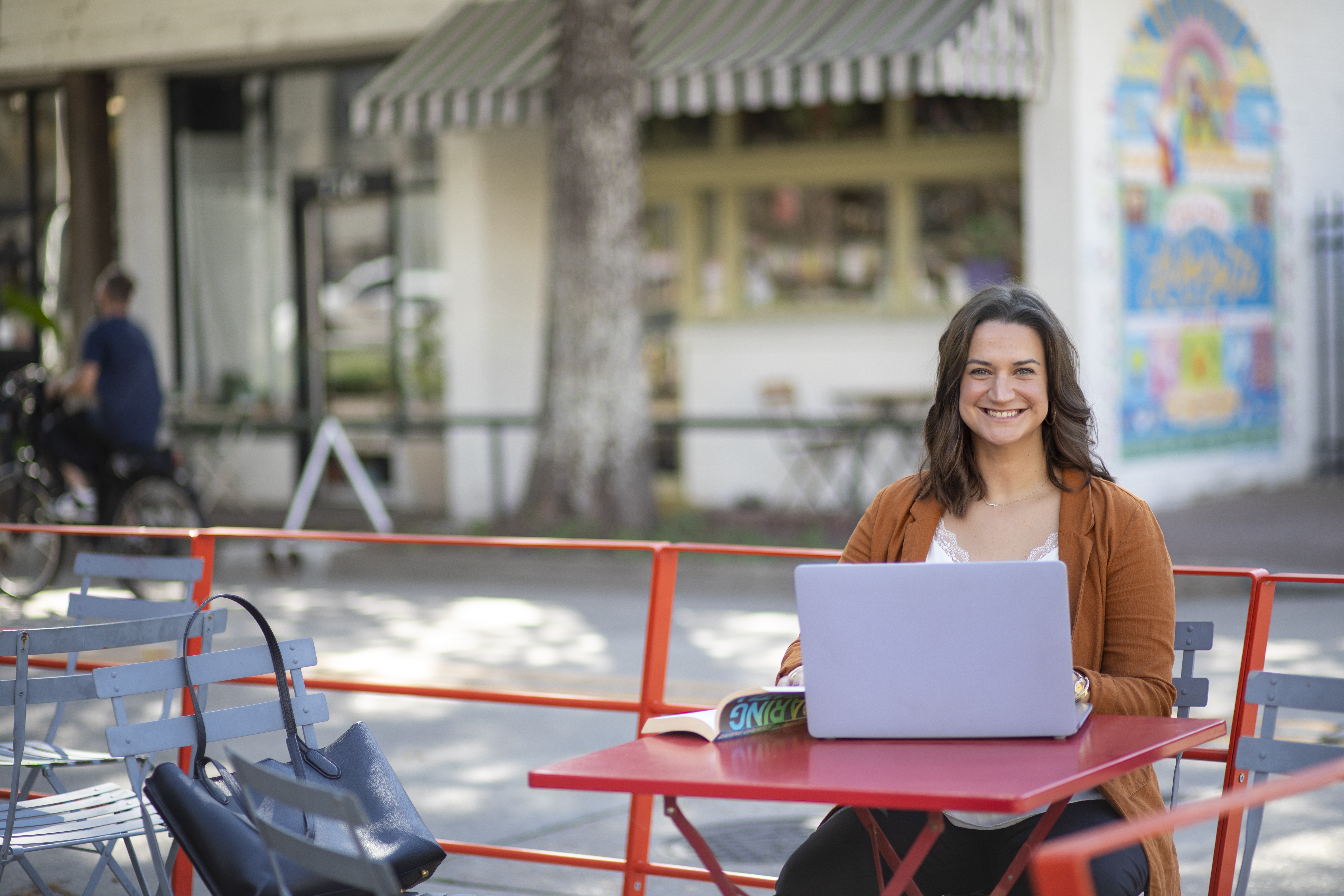 JoElle Judice is pictured outdoors at a red table with an open book and laptop. She has shoulder-length brown hair and is wearing a light brown blazer. 