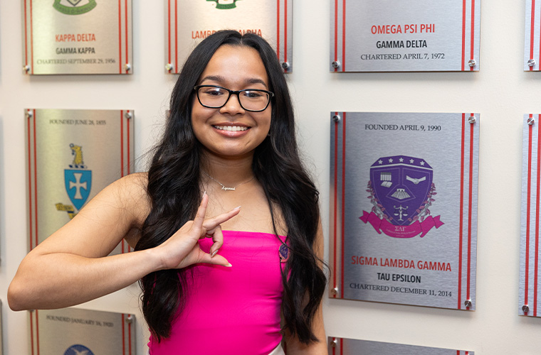 Sigma Lambda Gamma member standing next to a plaque for her sorority on UL Lafayette's Greek Wall.