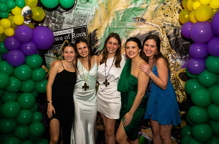 A group of UL Lafayette students posing together in formal dress surrounded my Mardi Gras decorations for the Krewe of Roux ball
