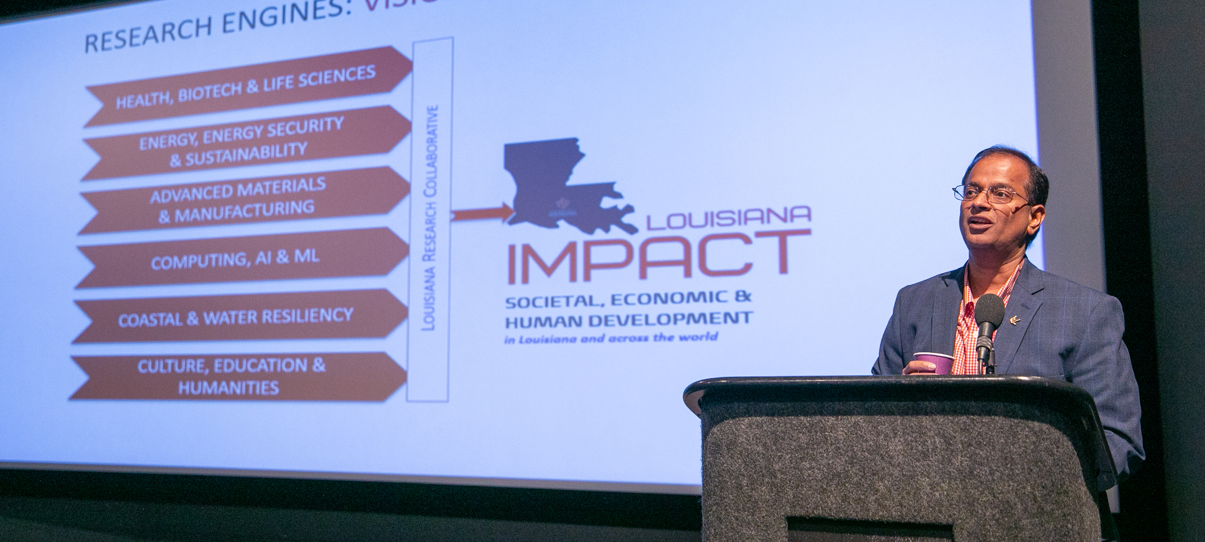 Dr. Ramesh Kolluru standing at a podium in front of a screen presenting on Louisiana Impact.