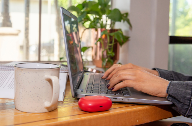 A woman's hands rest on the keyboard of an open laptop with a mug and a red earbud case in the foreground.
