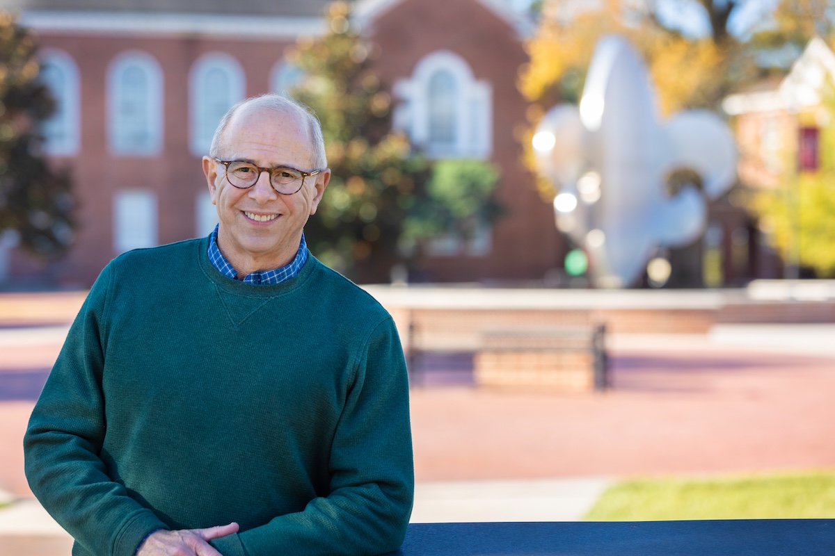A man wearing glasses and a green sweater smiles in front of the fleur de lis fountain in the UL Lafayette quad.