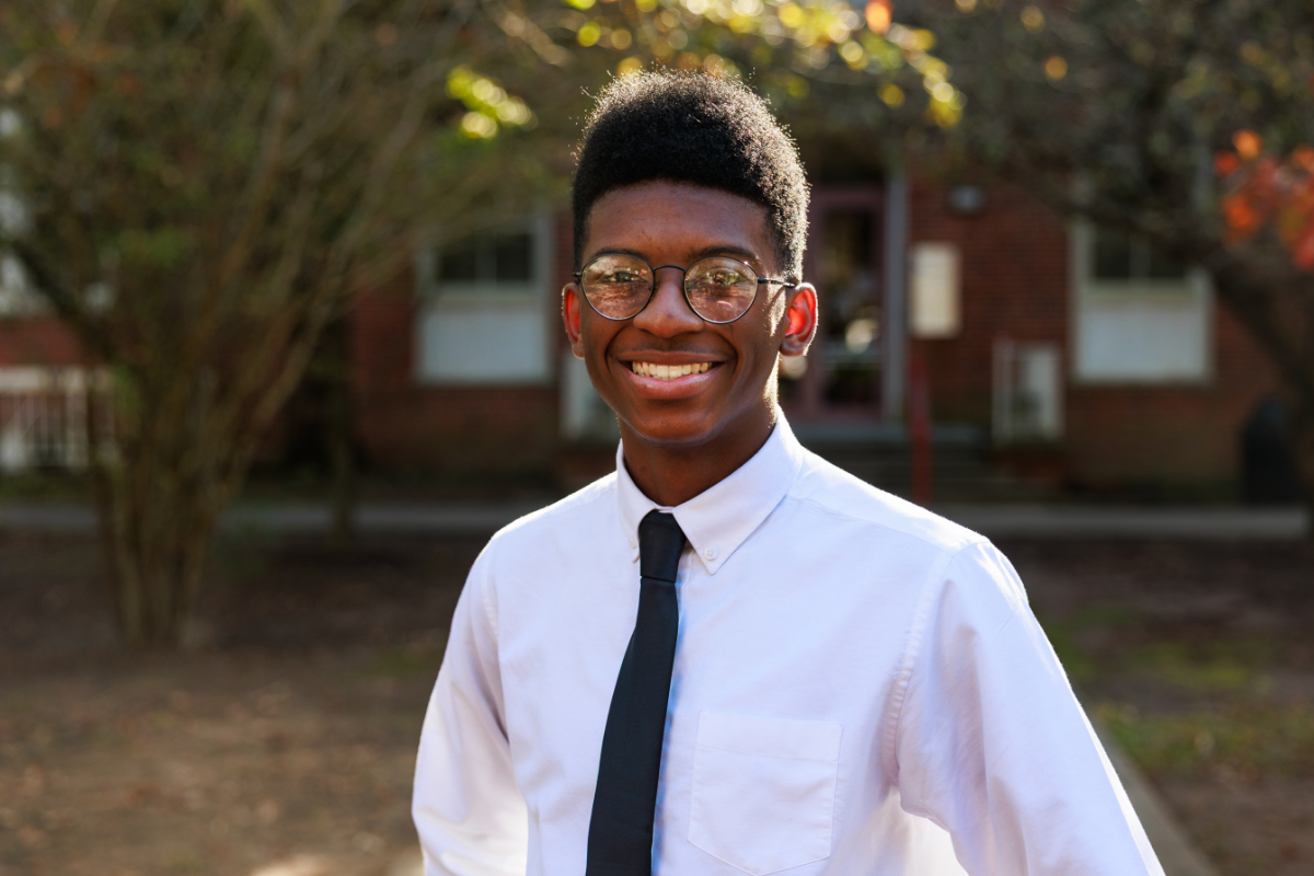 Portrait photograph of Xzayvion Gaye smiling wearing a white long sleeve button down dress shirt and a black tie. He also is wearing glasses standing in front of trees and a building.