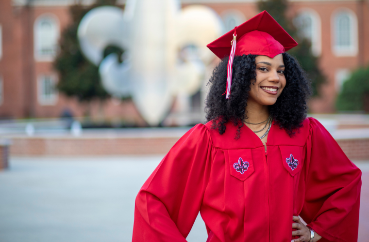 A young woman is pictured wearing a red graduation cap and gown in front of a metal fleur de lis sculpture in UL Lafayette's quad. 