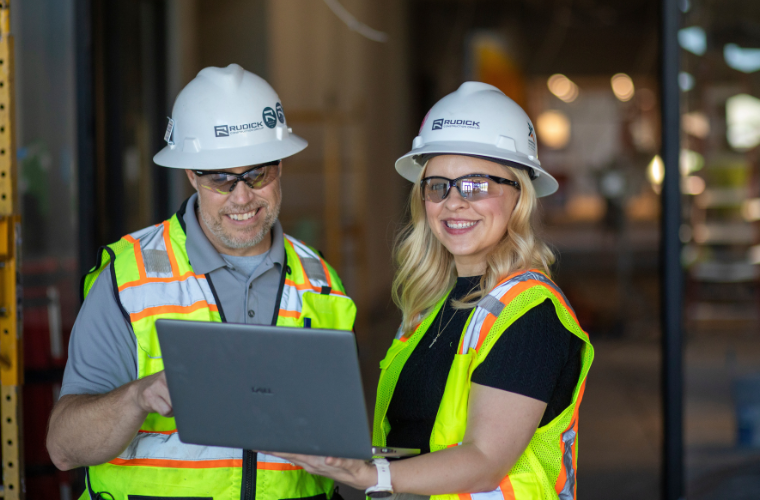 Online masters in project management grad collaborates on a worksite, wearing a neon vest and hard hat.