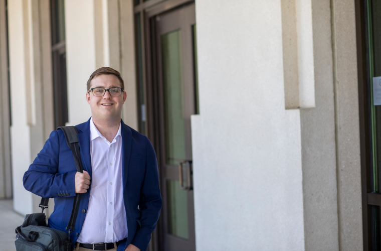 A man wearing glasses, a white shirt, and a blue suit jacket stands with a bag slung over their shoulder outside an office building.