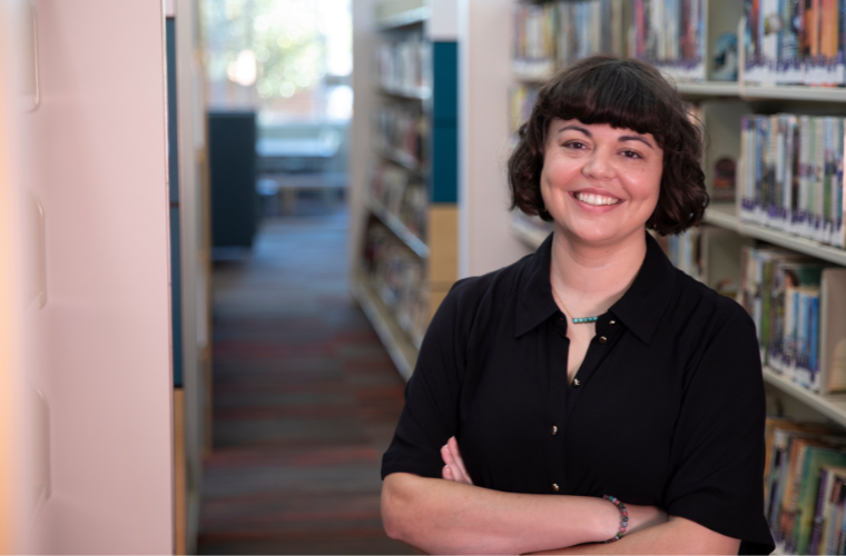 A woman with short dark hair smiles at the camera, standing with their arms crossed in a library aisle with bookshelves behind her.