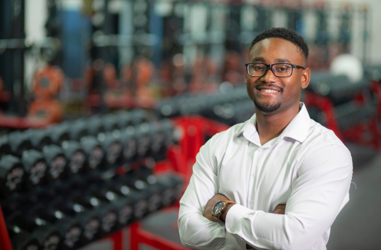 A man stands with his arms crossed near a free weight rack in a fitness center.