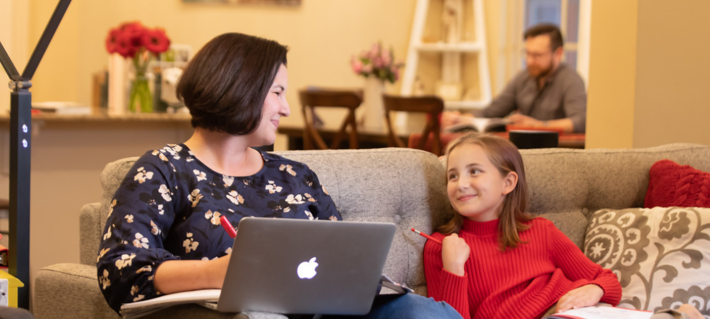 UL Lafayette online student working at a computer on the couch next to their child with their spouse in the background.