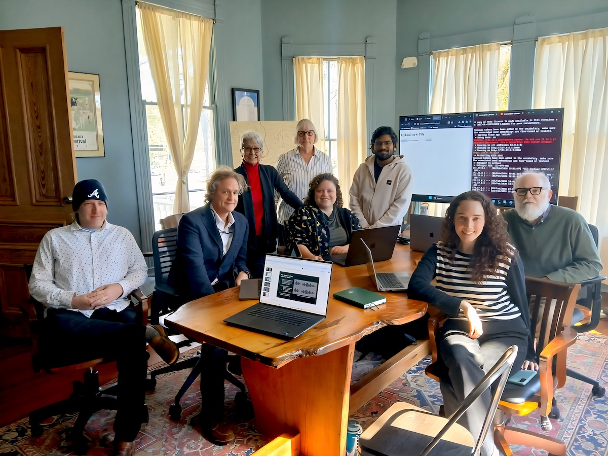 A group of eight people sit at a table in a conference room with a laptop and TV screen behind them.