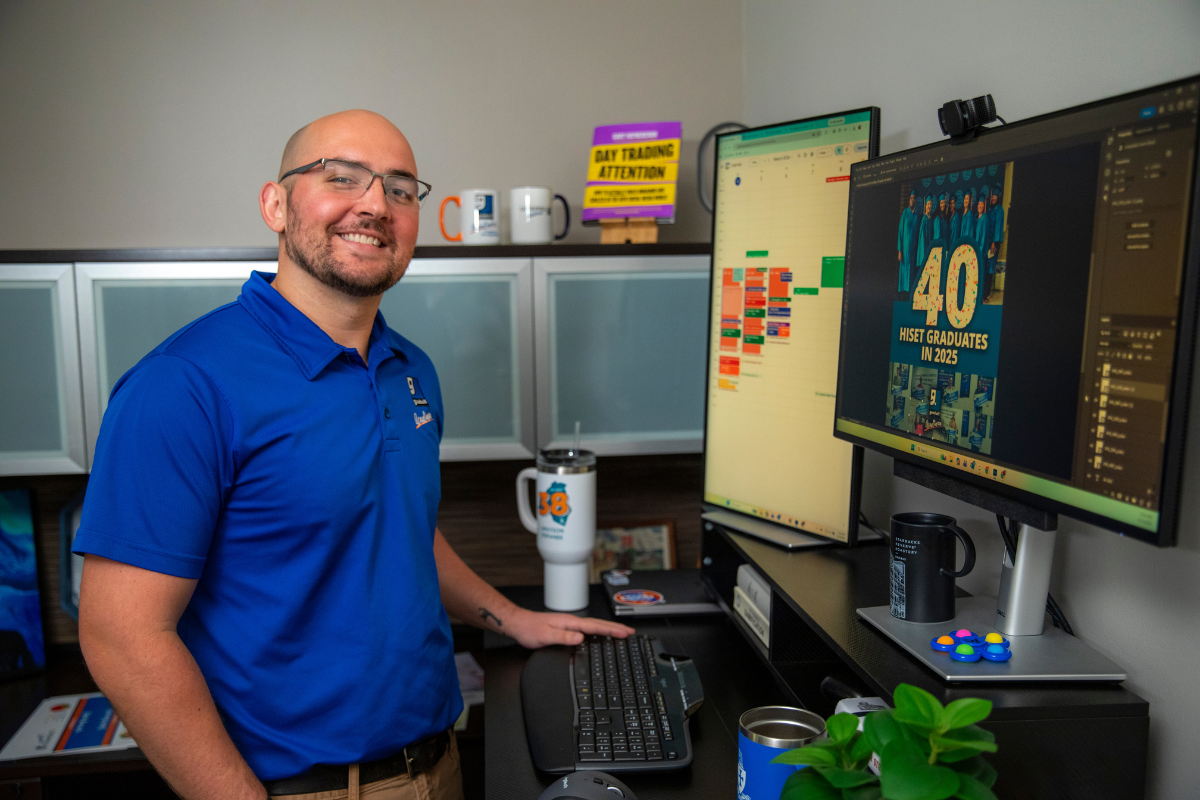Grayson Stepanek is pictured wearing a blue polo shirt and smiling at his office desk at Goodwill Acadiana.