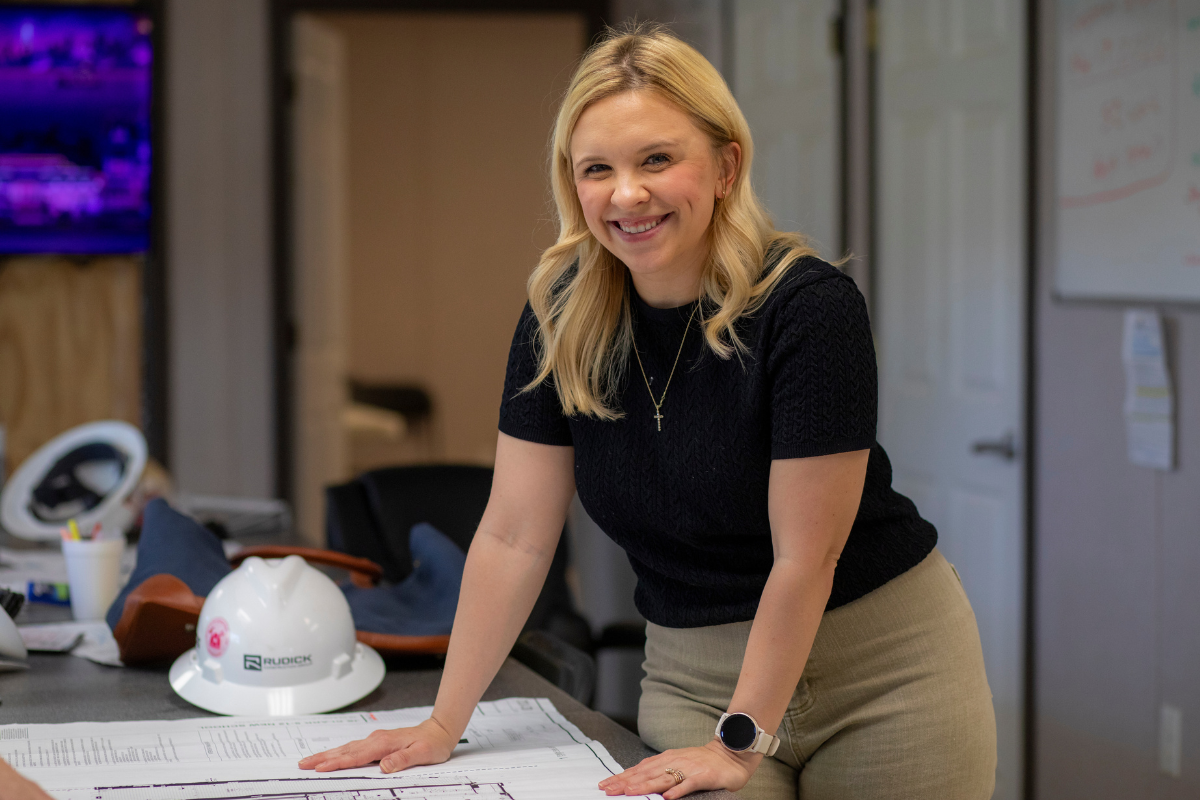 Jillian Blanchard smiles into the camera while leaning over a table laden with construction plans and a Rudick Construction Group hard hat.
