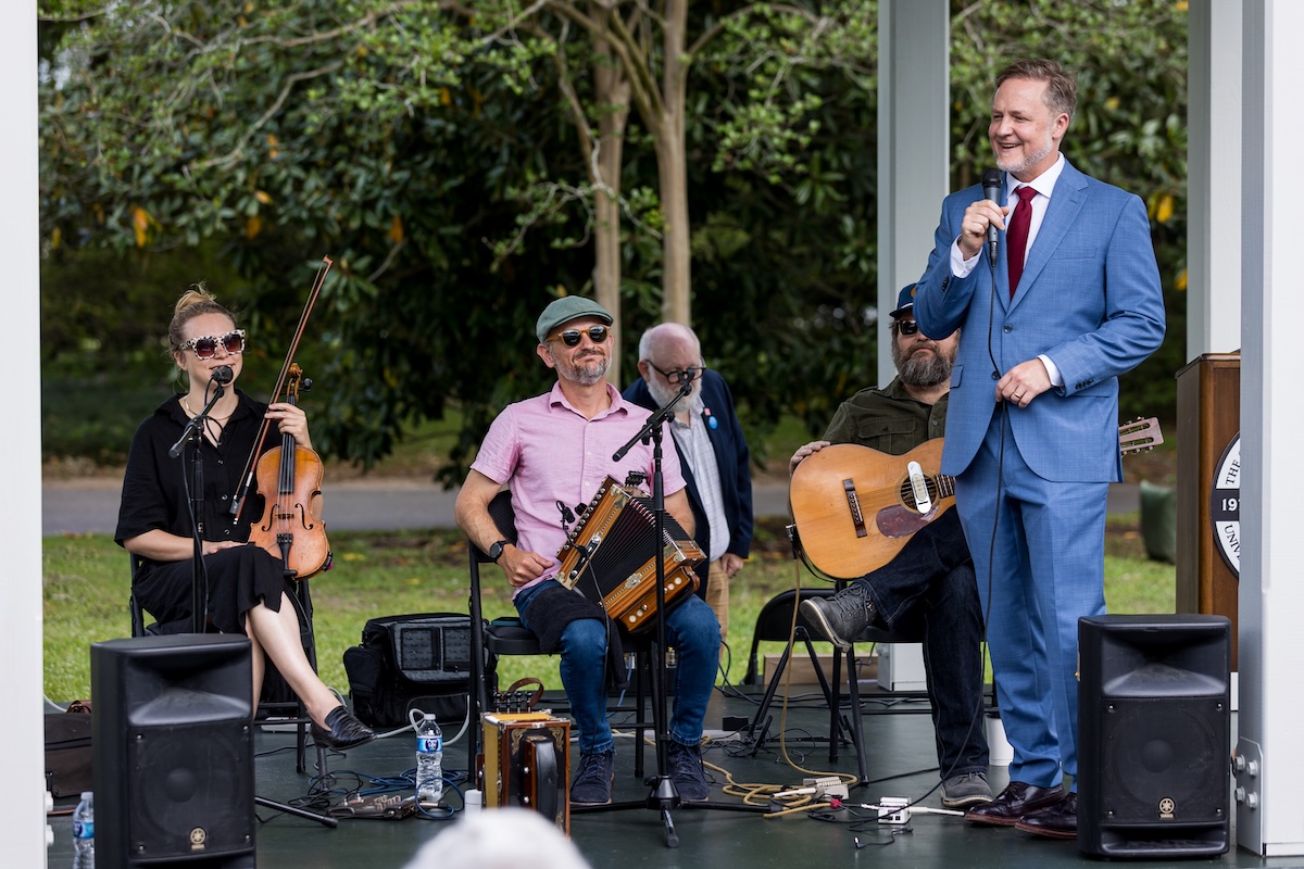 A man in a suit holds a microphone while standing in an outdoor pavilion in front of a three-piece Cajun band.
