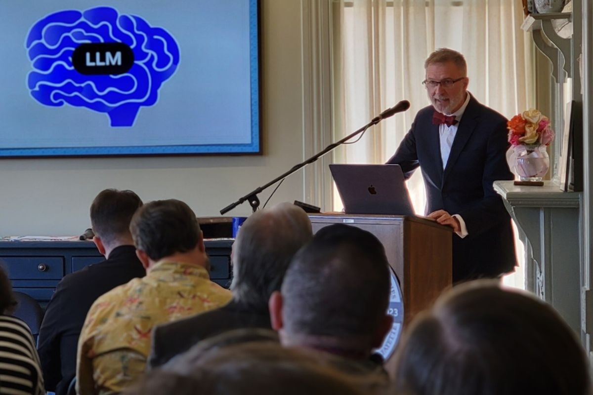Man in suit and bowtie stands behind a lecturn with a screen behind him featuring an illustration of a brain and the letters LLM.
