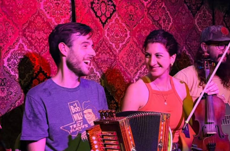 A man holds an accordion next to a woman holding a fiddle, smiling