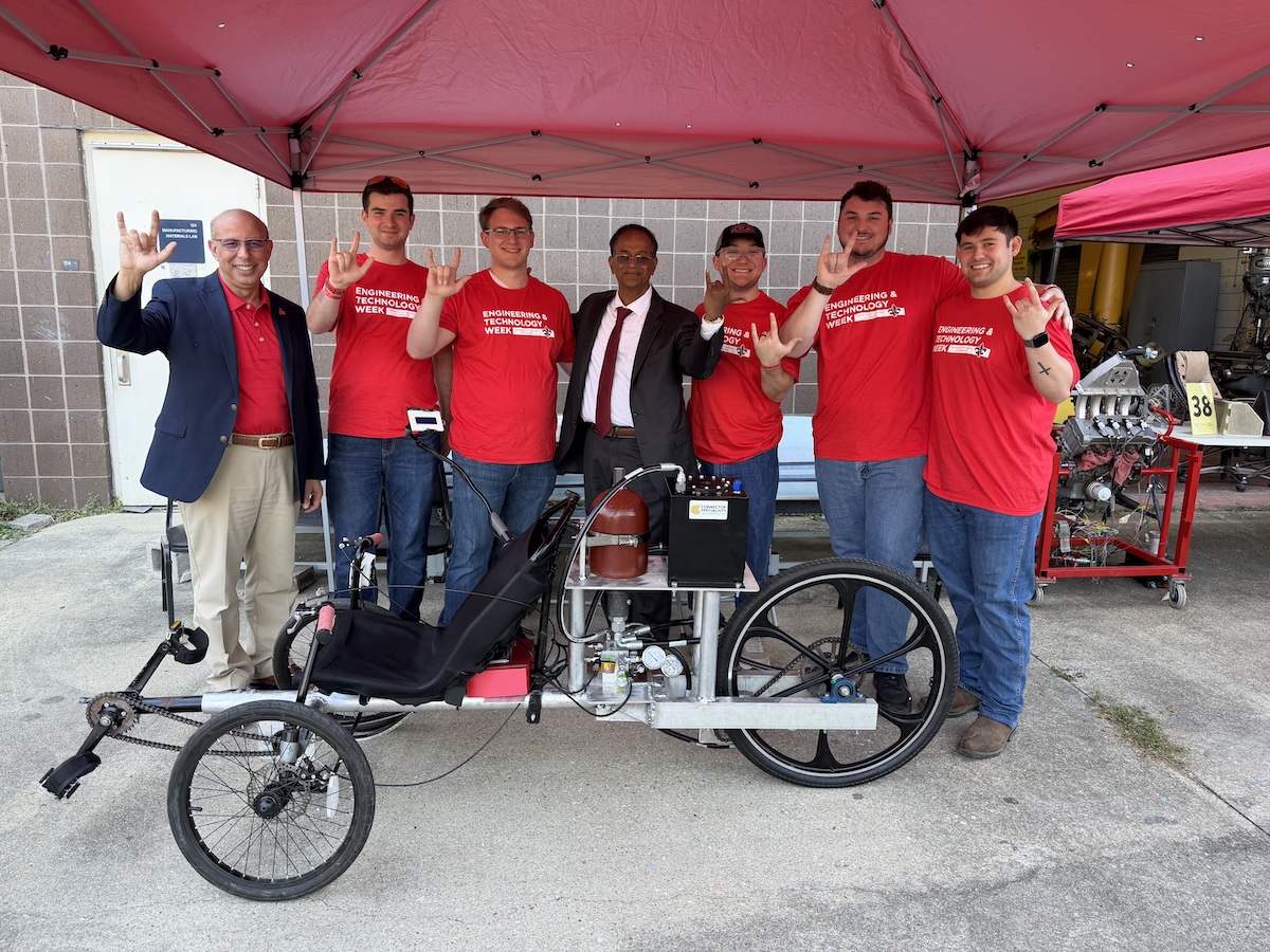 Seven people stand in front of a hydraulic vehicle.