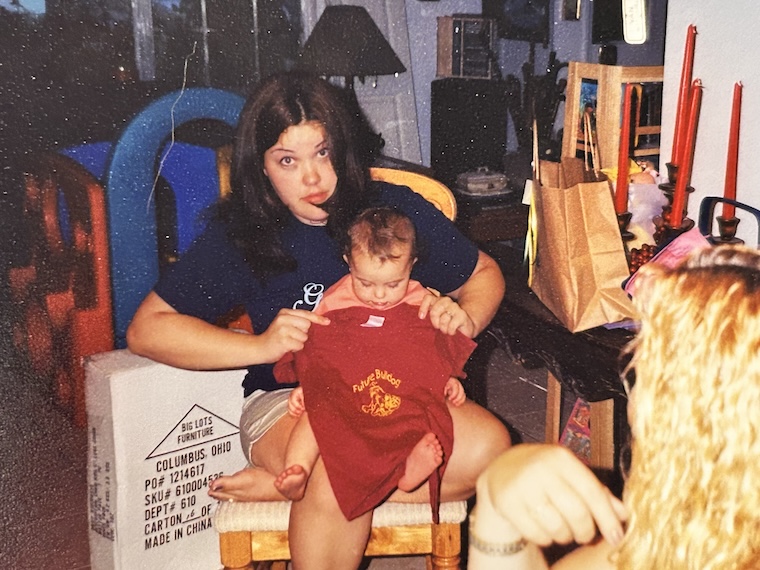 A mother holds a one-year-old baby on her lap while holding a red shirt up to her baby that says "Future Bulldog."