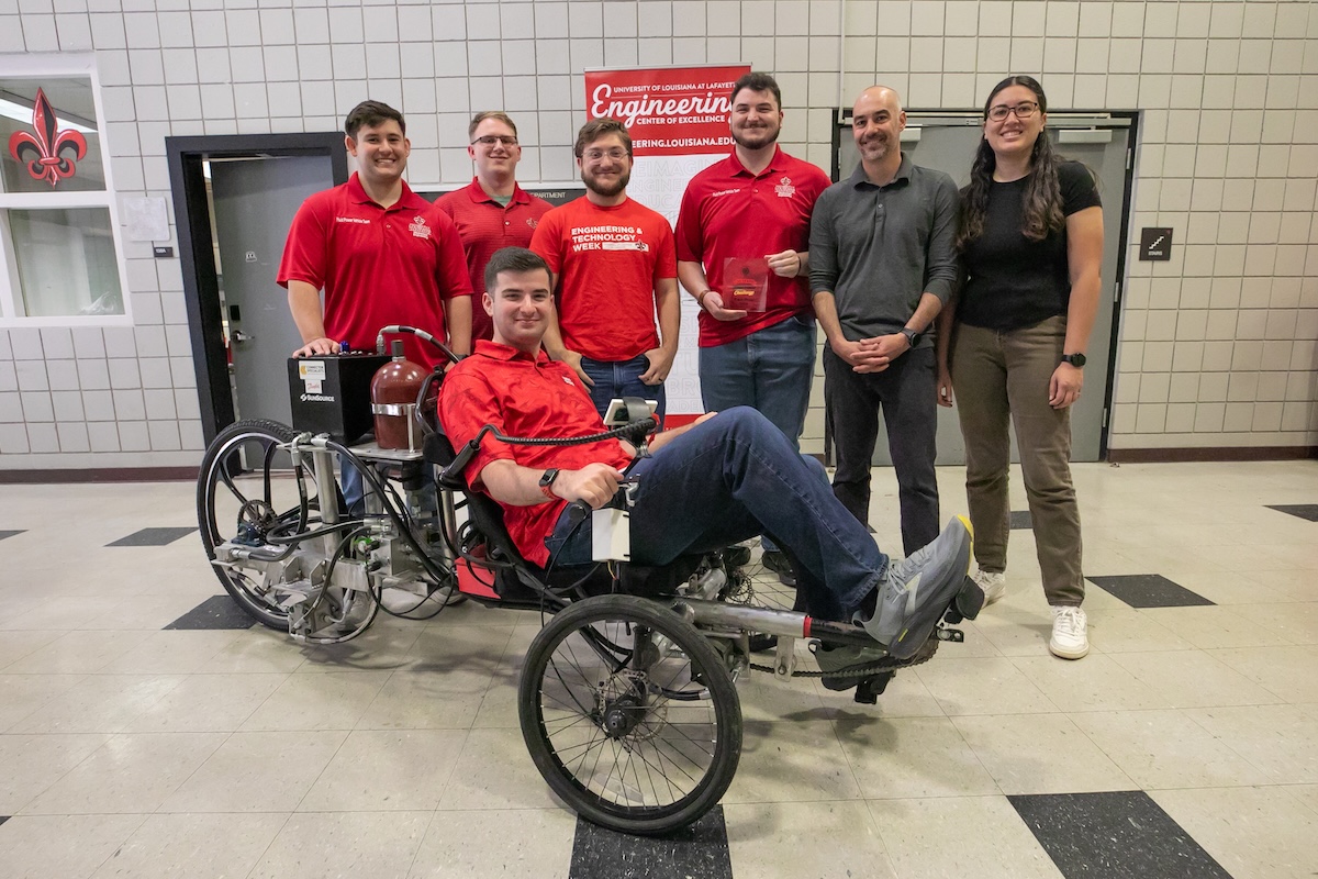 Six people stand for a group photo with one person sitting in a tricycle holding a trophy.