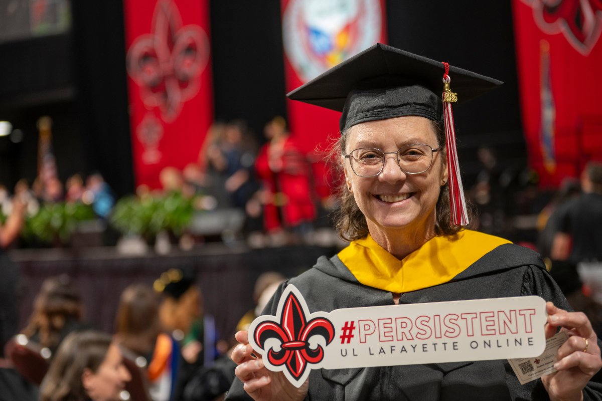 Karen LeDoux is pictured during commencement ceremonies wearing UL Lafayette's black master's cap and gown and holding a sign that reads: #PERSISTENT UL Lafayette Online.