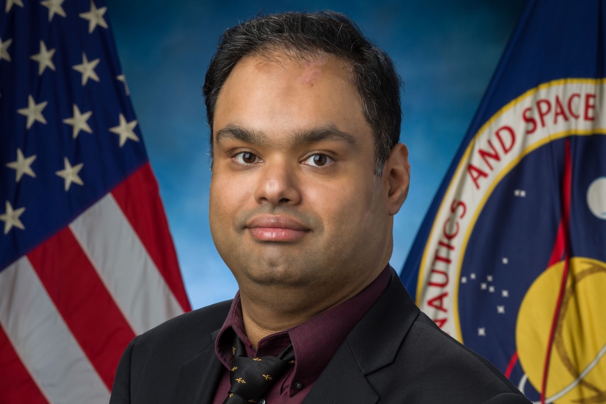 A headshot of a man in front of an American flag and a NASA flag.