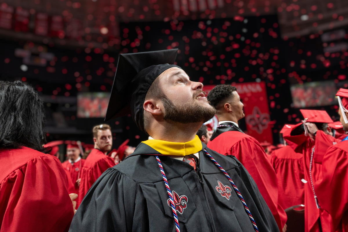 Online master's graduate wears a black cap and gown, gazing up as red confetti falls.