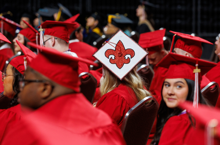 UL Lafayette graduation cap decorated with a fleur de lis in a sea of graduating students in red graduation robes.