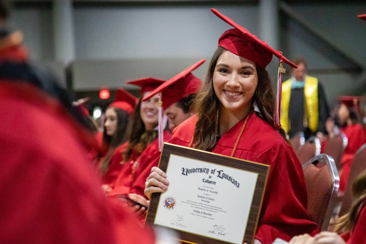 Graduate of the online kinesiology degree program smiles in a cap and gown, holding her newly earned bachelor's degree plaque.